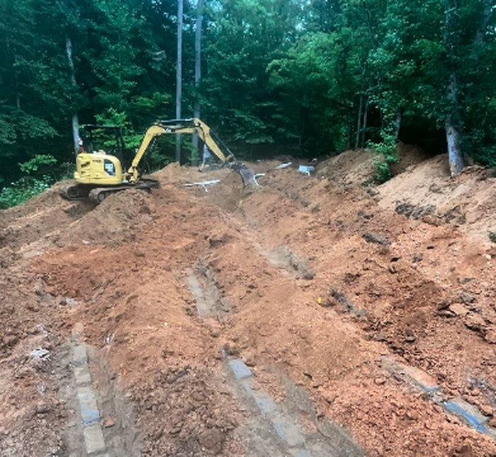 Excavator trenching a septic drain line across a wooded Troutman lot